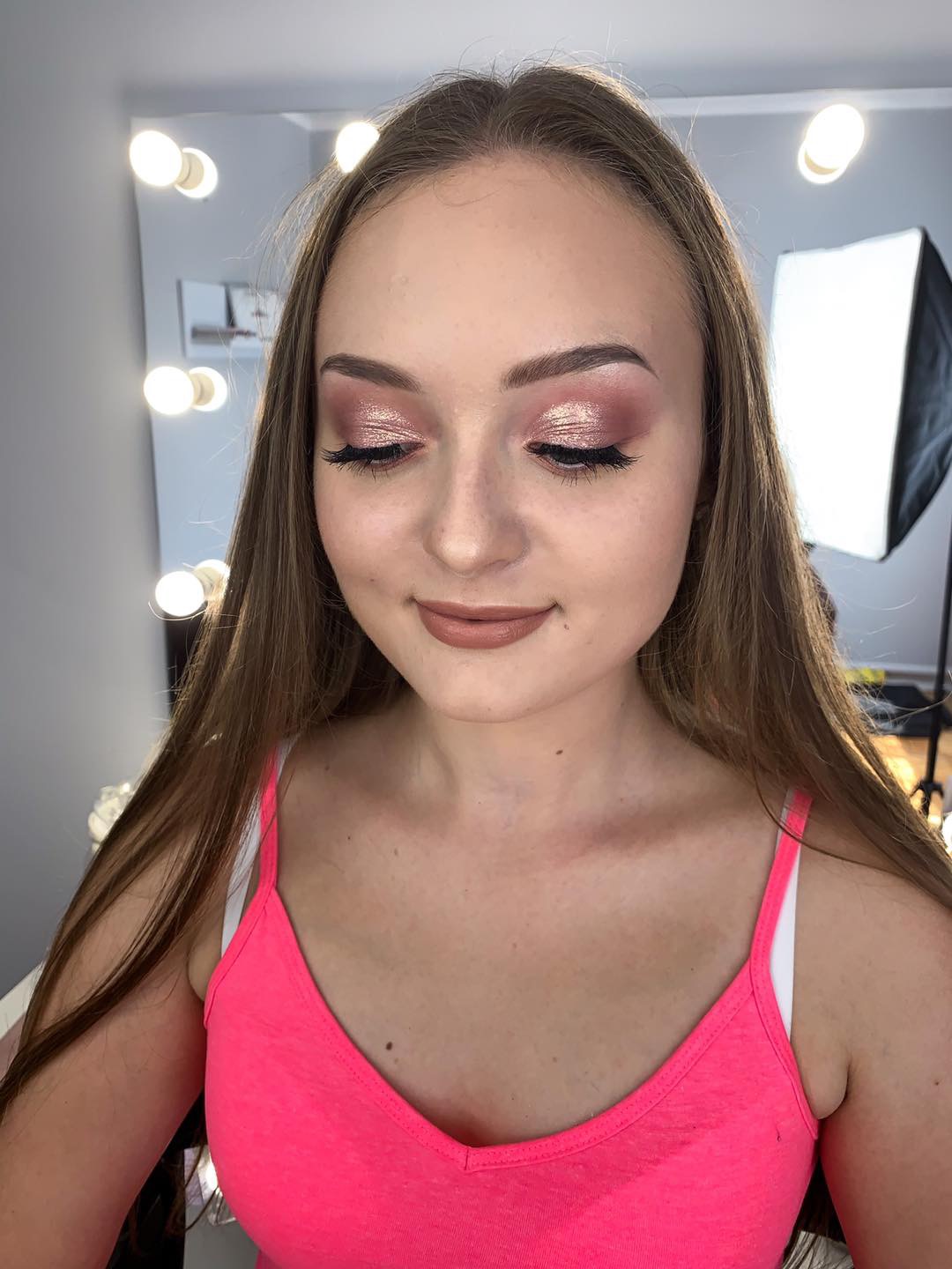 Portret kobiety z błyszczącym, różowym cieniem na powiekach, długimi sztucznymi rzęsami i brązową pomadką, pozującej przed lustrem z oświetleniem i softboxem.