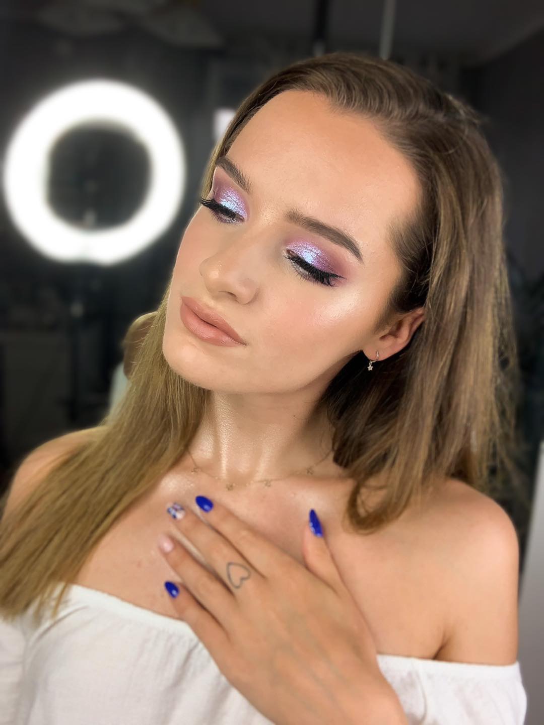 Portret kobiety z zamkniętymi oczami, prezentującej opalizujący, fioletowo-różowy cień na powiekach i błyszczące usta, z dłonią ozdobioną niebieskim manicure i tatuażem serca, na tle studyjnego...