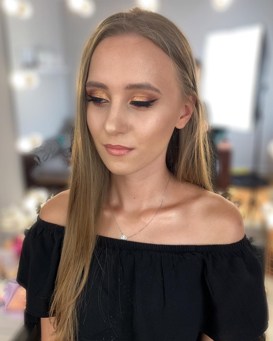 Portret kobiety z profesjonalnie wykonanym, złotym cieniowaniem powiek i czarną kreską, uzupełnionym delikatnym błyszczykiem na ustach i srebrnym naszyjnikiem z małą zawieszką.