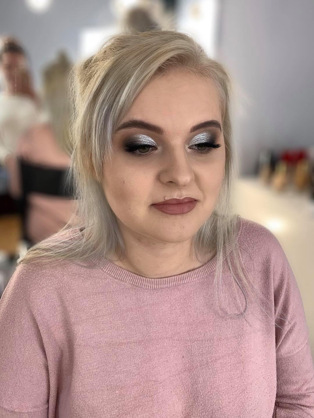 Portret kobiety z platynowymi włosami i srebrzystym cieniem na powiekach, z delikatnym różem na ustach, ubrana w różowy sweter na rozmytym tle salonu kosmetycznego.
