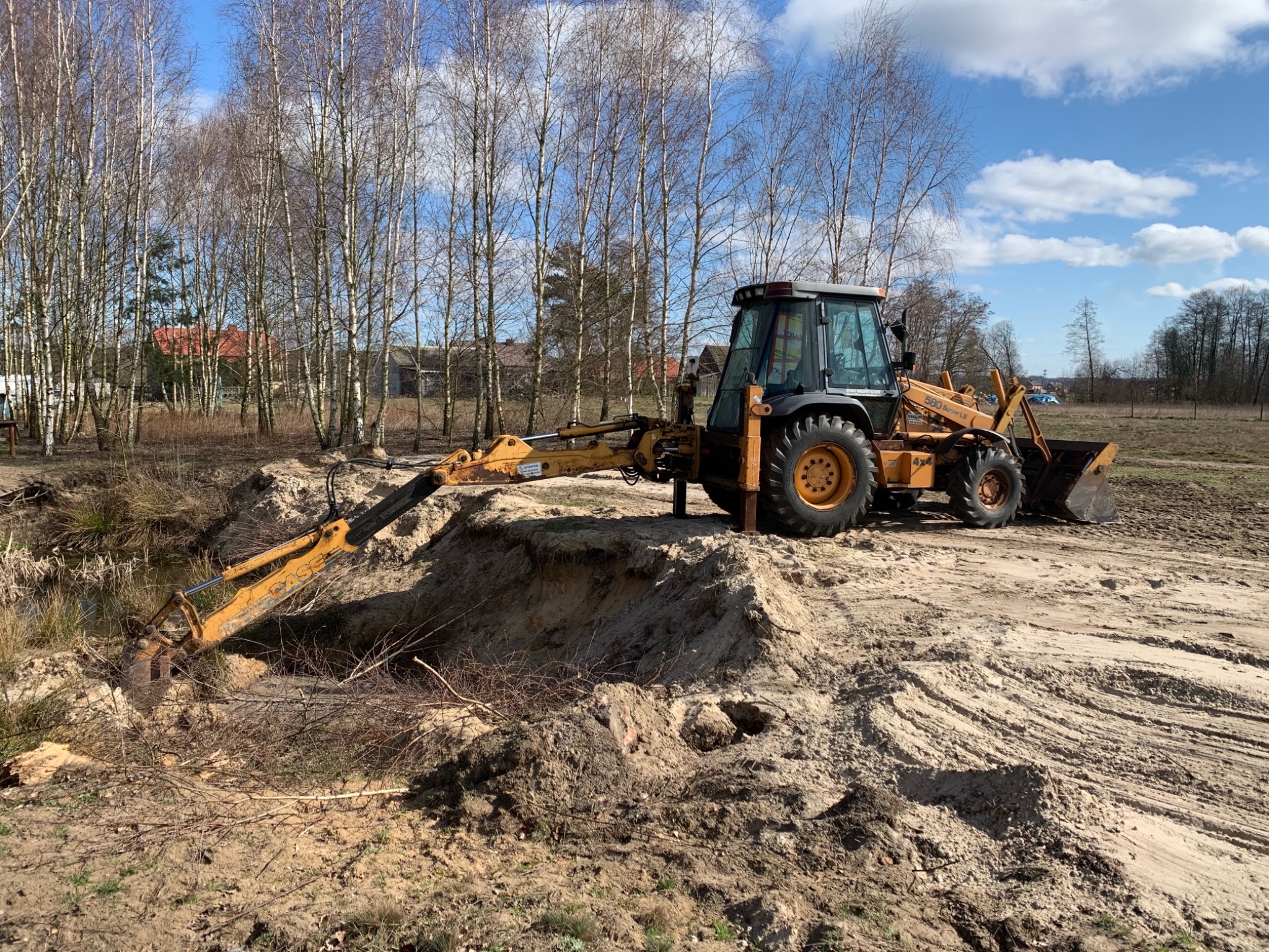 Żółta koparko-ładowarka CASE 580 Super LE podczas pogłębiania rowu melioracyjnego na piaszczystym terenie z młodymi brzozami w tle.