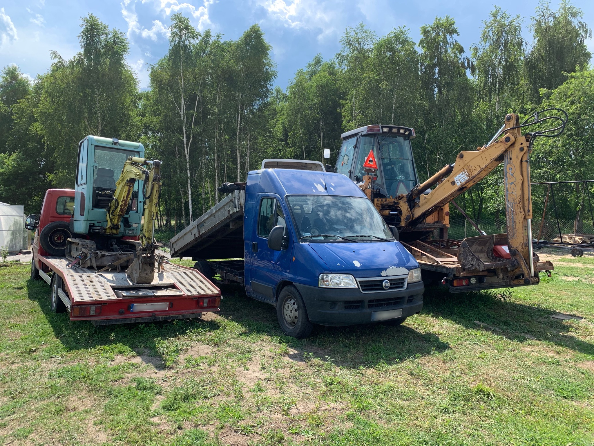 Transport maszyn budowlanych: mała koparka na czerwonej lawecie, niebieski bus z platformą i koparko-ładowarka w tle, na zielonej trawie, drzewa w tle.