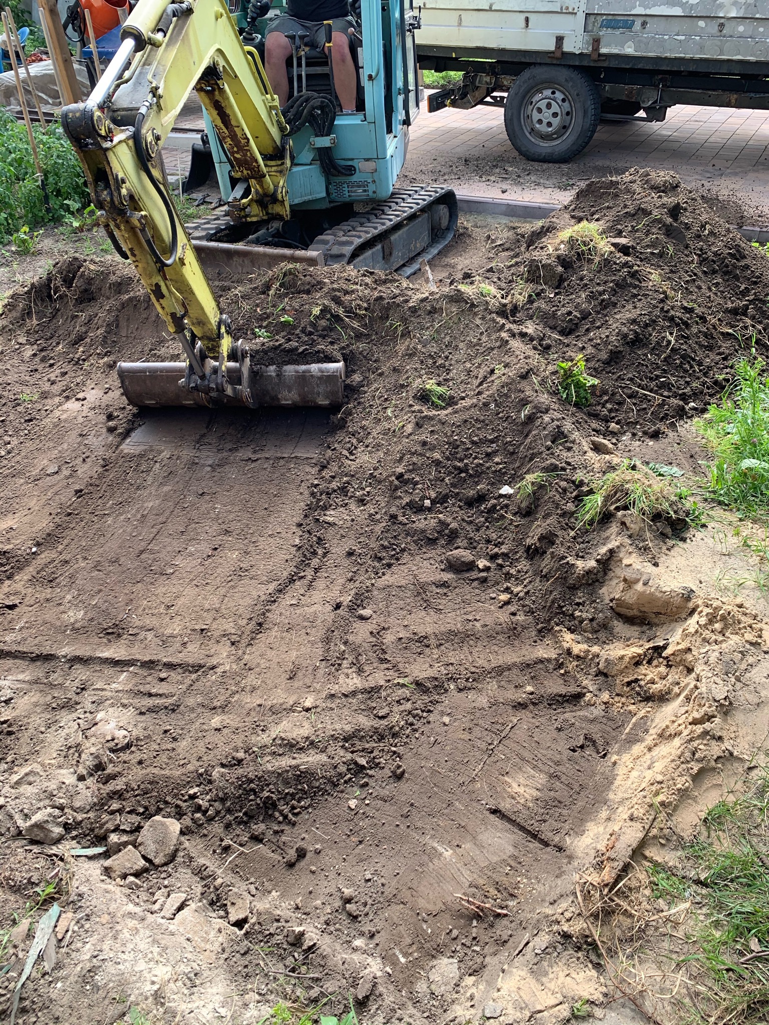 Żółto-błękitna mini koparka podczas prac ziemnych na działce, widoczny wykopany obszar z wyraźnymi śladami łyżki koparki, pryzma ziemi obok oraz fragment samochodu dostawczego w tle.