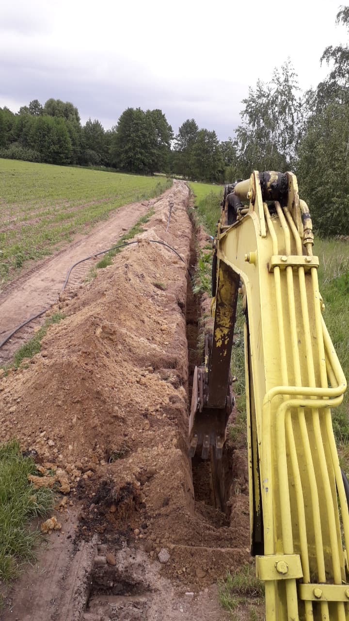 Żółta koparka wykonuje wąski wykop w ziemi na tle pola i drzew, widoczny kabel wzdłuż wykopu.
