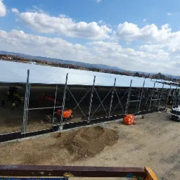 Budowa hali stalowej z widocznymi maszynami budowlanymi, w tle zabudowa mieszkalna i góry, częściowo zamontowana konstrukcja dachu.