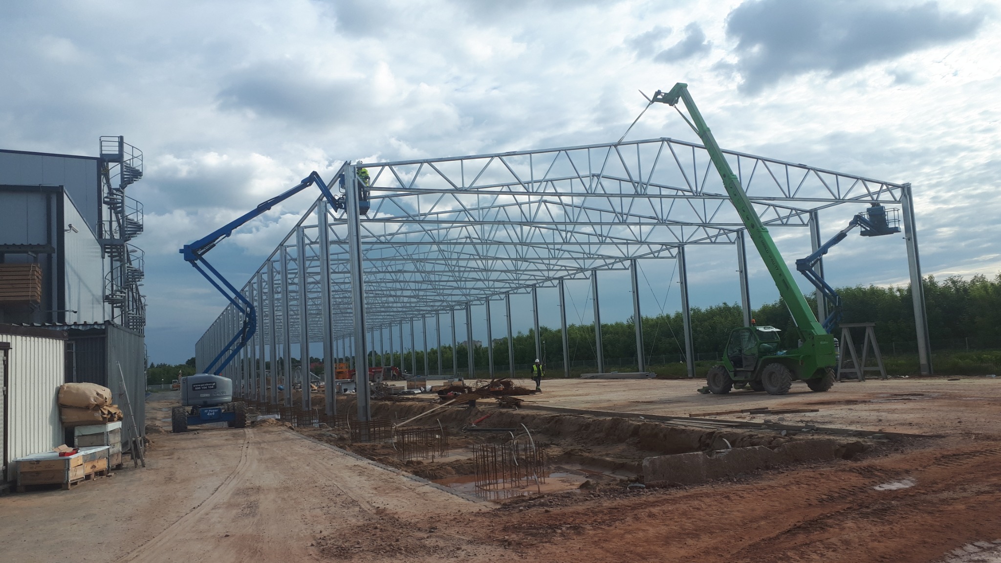 Wznoszenie szkieletu stalowej hali z użyciem podnośników koszowych i teleskopowych, widoczne fundamenty i przygotowany teren.