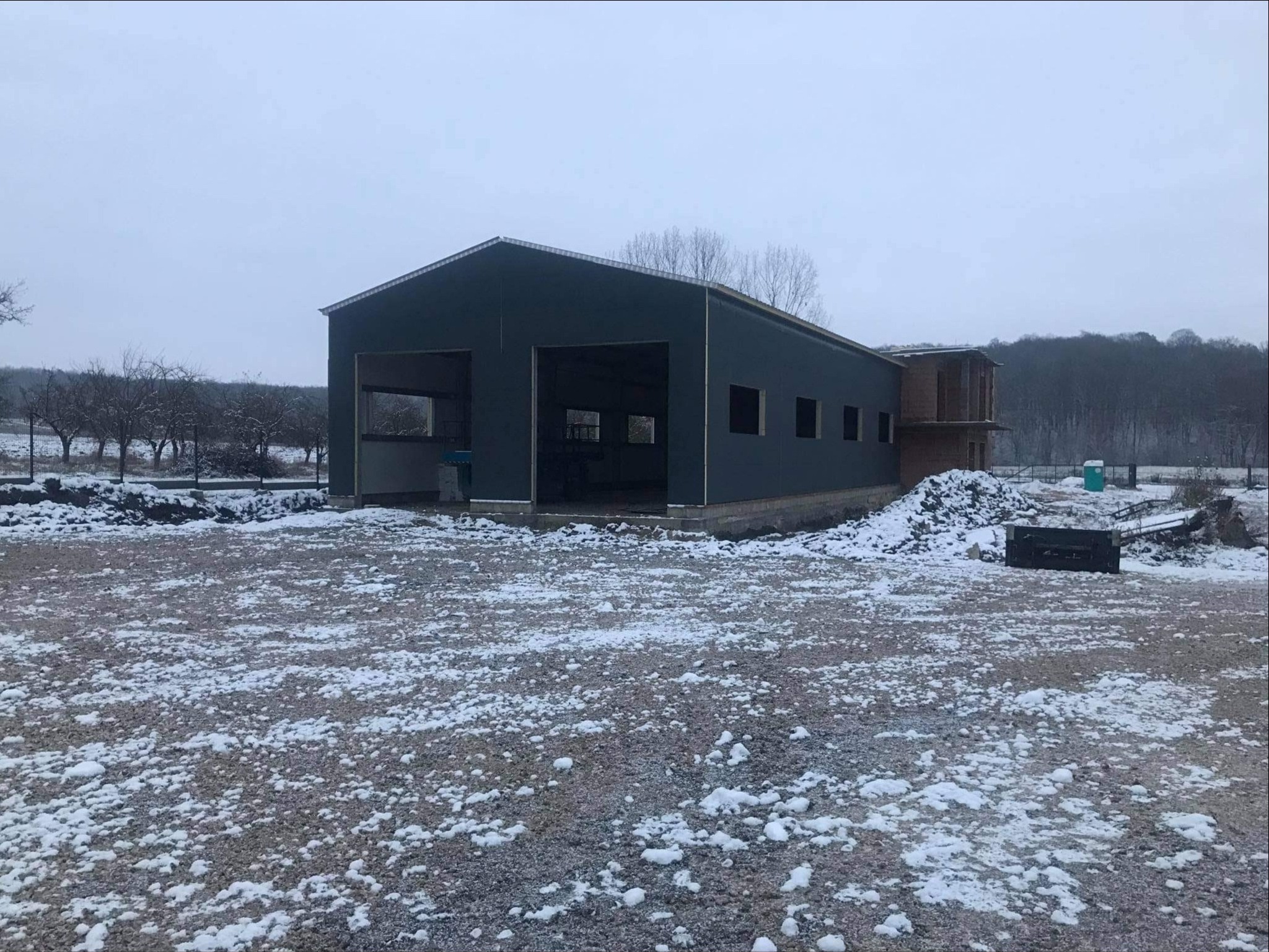 Szara hala stalowa w trakcie budowy zimą, widoczne niezamknięte otwory okienne i drzwiowe, fundamenty oraz częściowo ośnieżony teren budowy z widocznym drzewostanem w tle.