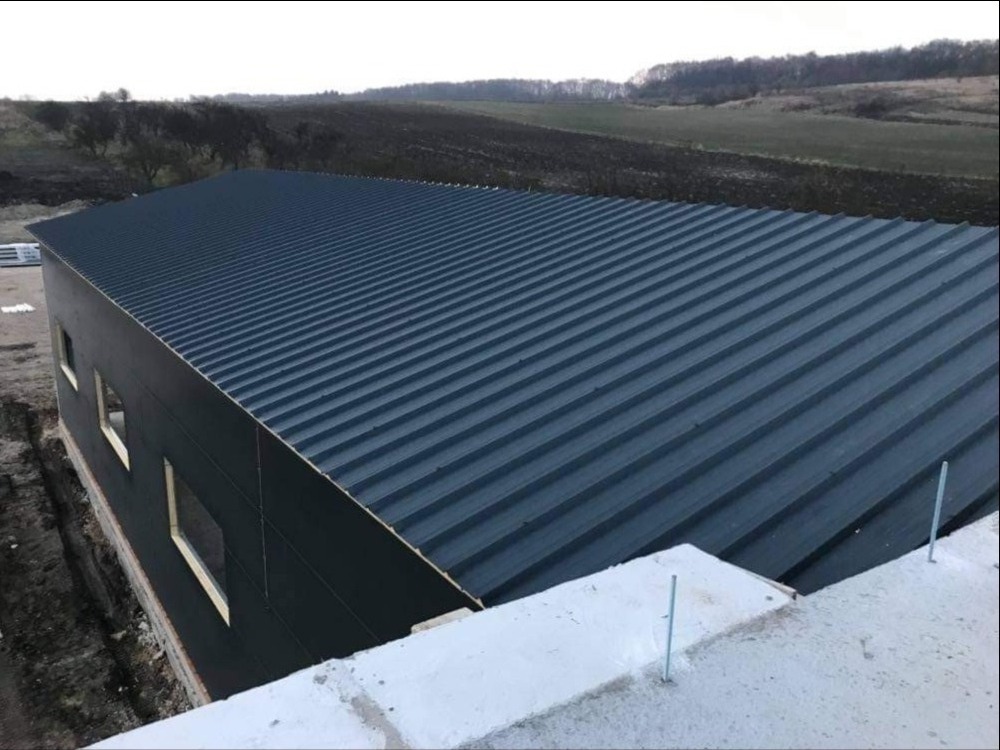 Nowo wybudowana hala z ciemnymi panelami warstwowymi i dachem z blachy trapezowej na tle zaoranego pola i lasu.