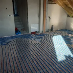 Niebieskie rurki ogrzewania podłogowego ułożone w pętle na siatce, częściowo oświetlone przez wpadające światło słoneczne, widoczne surowe ściany i drewniana konstrukcja dachu poddasza.