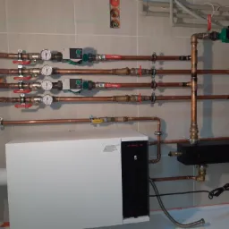 Instalacja grzewcza z białym kotłem, miedzianymi rurami, zaworami Ferro i manometrami na tle płytek ceramicznych. Widoczne również pompy obiegowe i czarna skrzynka rozdzielcza.