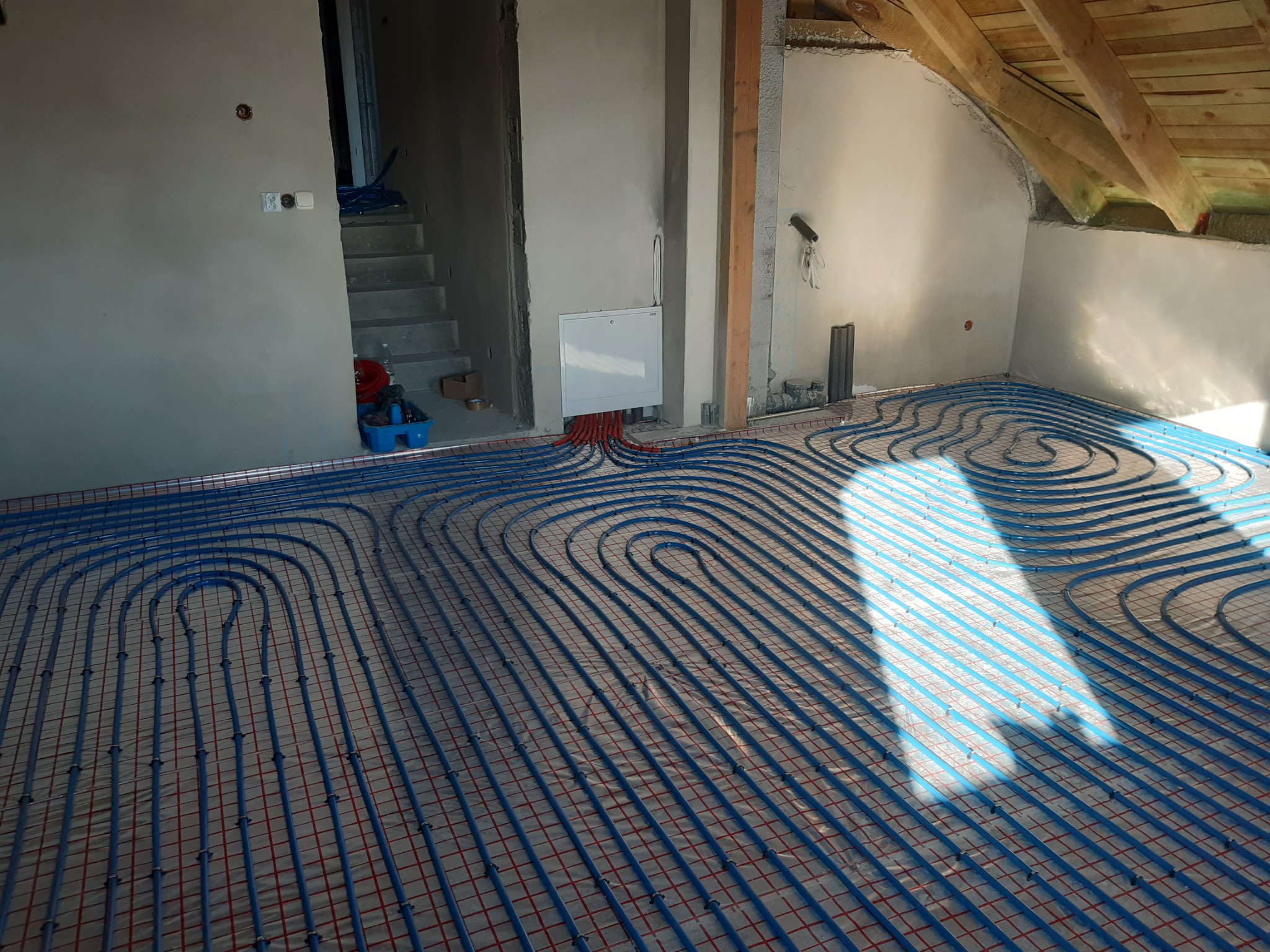Niebieskie rurki ogrzewania podłogowego ułożone w pętle na siatce, częściowo oświetlone przez wpadające światło słoneczne, widoczne surowe ściany i drewniana konstrukcja dachu poddasza.