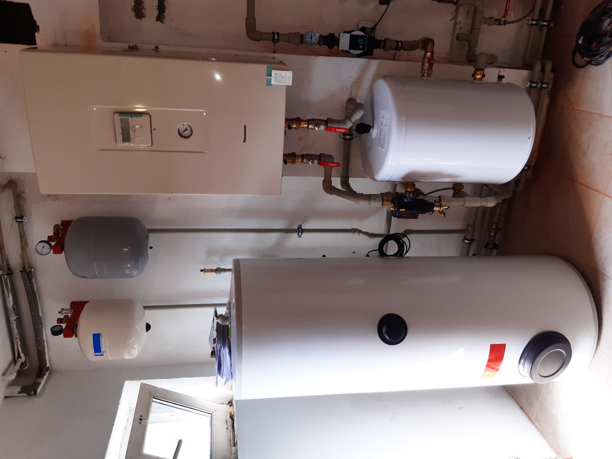 Zamontowany zestaw centralnego ogrzewania w kotłowni: wiszący piec gazowy Panasonic, dwa białe zasobniki wody i szary zbiornik wyrównawczy, połączone rurami z zaworami.