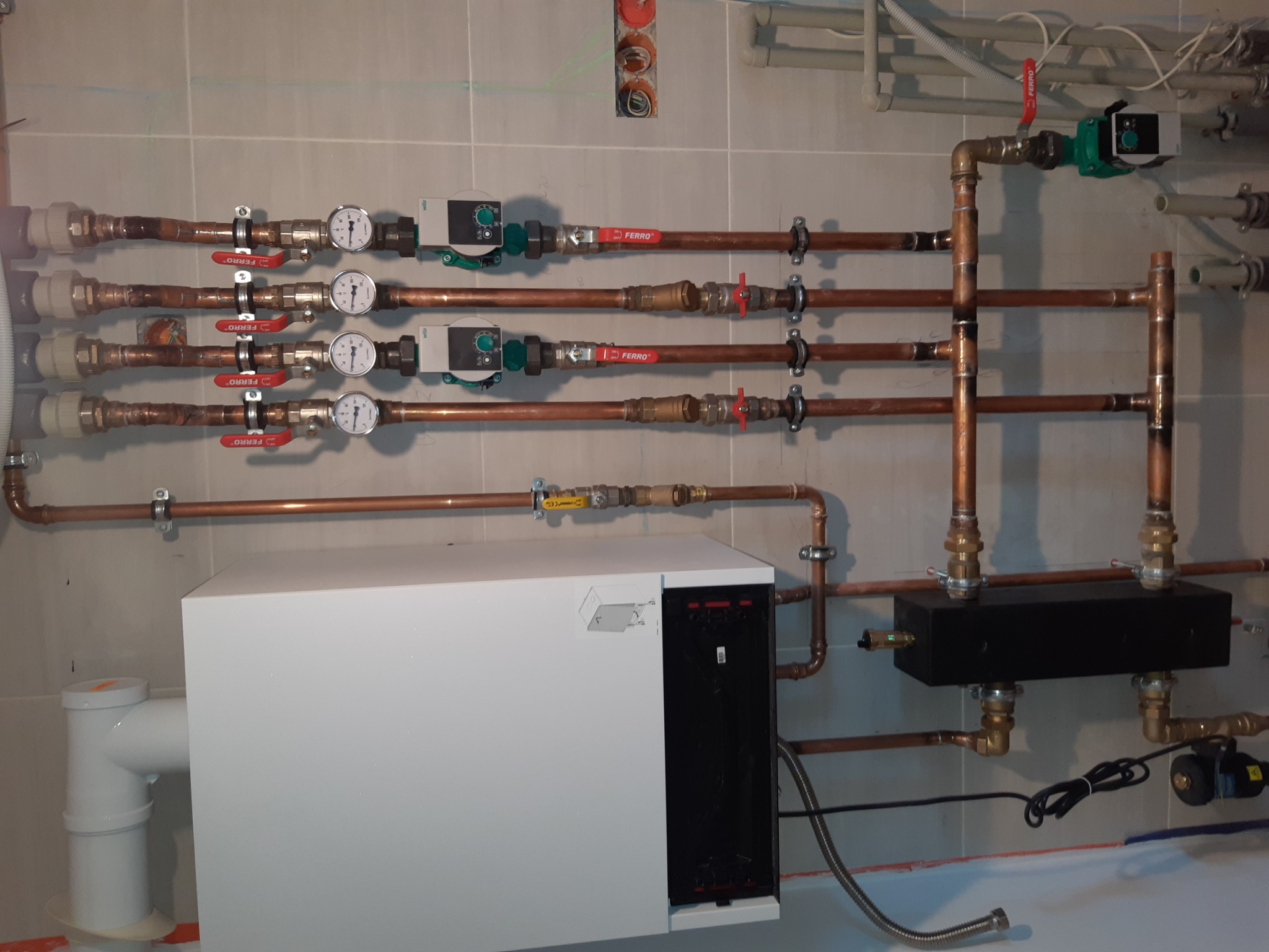 Instalacja grzewcza z białym kotłem, miedzianymi rurami, zaworami Ferro i manometrami na tle płytek ceramicznych. Widoczne również pompy obiegowe i czarna skrzynka rozdzielcza.