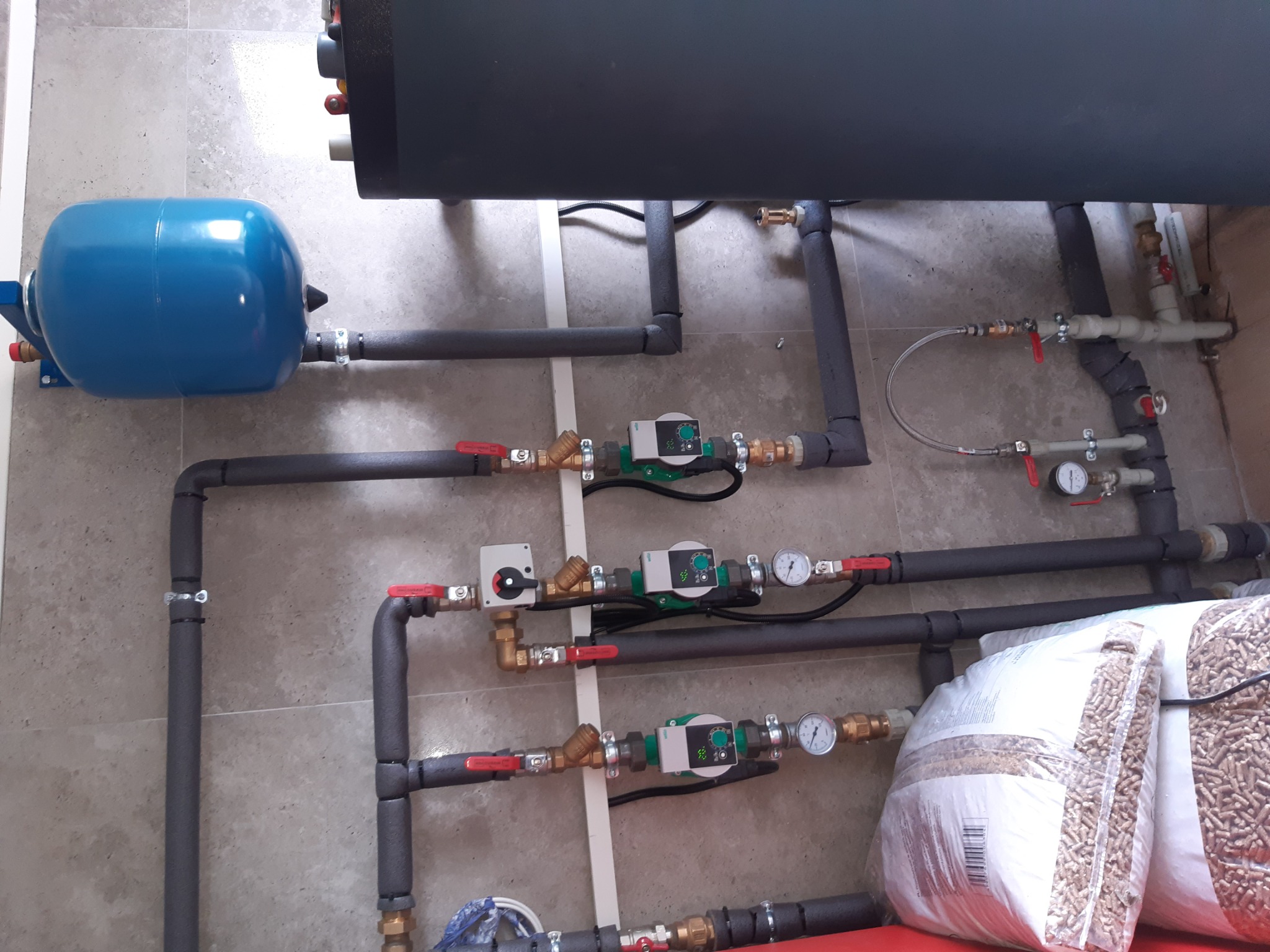 Instalacja kotłowni z zasobnikiem, pompami obiegowymi, zaworami i workami pelletu na podłodze z płytek.