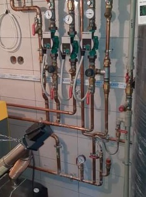 Instalacja centralnego ogrzewania z trzema pompami obiegowymi, manometrami i zaworami, widoczne miedziane rury i elementy montażowe na tle kafelkowanej ściany.