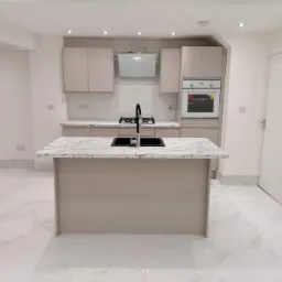 Nowoczesna kuchnia w odcieniach beżu i bieli z wyspą kuchenną z marmurowym blatem i czarną armaturą, oświetlona punktowymi lampami sufitowymi, z białymi płytkami na ścianie nad blatem.