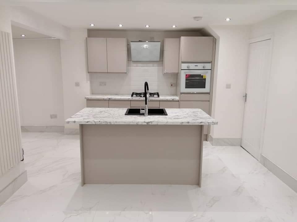 Nowoczesna kuchnia w odcieniach beżu i bieli z wyspą kuchenną z marmurowym blatem i czarną armaturą, oświetlona punktowymi lampami sufitowymi, z białymi płytkami na ścianie nad blatem.