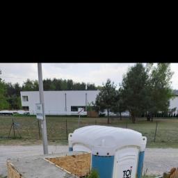 Seb-Bud - Tymczasowa toaleta na placu budowy w Warszawie, obok widoczna betonowa konstrukcja w drewnianym szalunku, w tle nowoczesny budynek i drzewa.