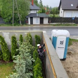 Seb-Bud - Pracownik w roboczym ubraniu, w trakcie wylewania betonu na mur oporowy, widoczna przenośna toaleta Toi Toi, oraz rząd tujek i świerków posadzonych wzdłuż muru.