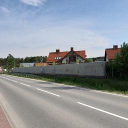 Seb-Bud - Długi, świeżo otynkowany mur oddzielający posesję od drogi, widoczny pracownik w jasnym ubraniu wykonujący drobne poprawki, w tle domy z czerwoną dachówką i błękitne niebo.