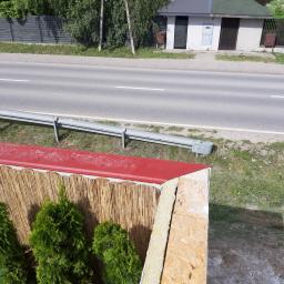 Seb-Bud - Fragment ogrodzenia z płyty OSB, ociepleniem i czerwoną obróbką blacharską, z widokiem na ulicę i szpaler tuj.