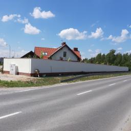 Seb-Bud - Mężczyzna pochylony przy długim, białym murze granicznym, prawdopodobnie wykonuje prace wykończeniowe, widoczna puszka z farbą i wałek, w tle dom z czerwoną dachówką i błękitne niebo z chmurami.