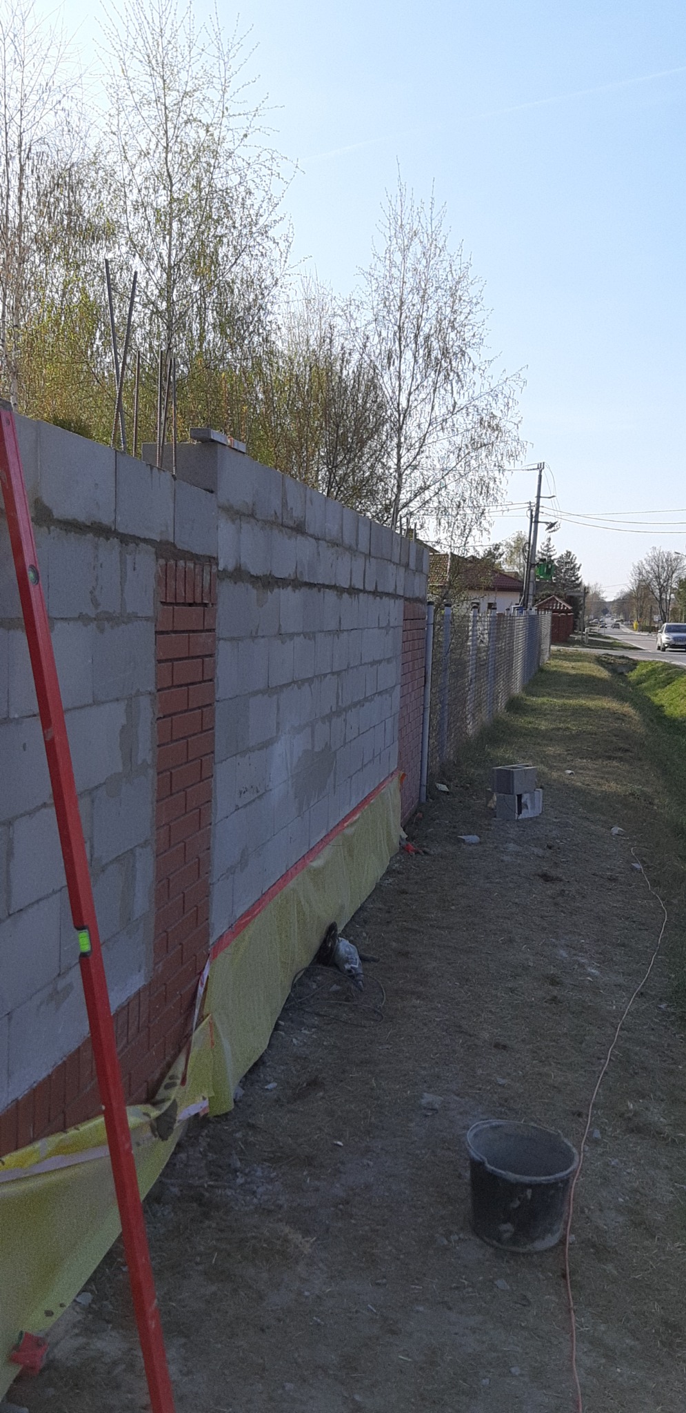 Wznoszenie ogrodzenia z bloczków betonowych z elementami z cegły, widoczny poziomica, wiadro, folia ochronna i szlifierka kątowa.