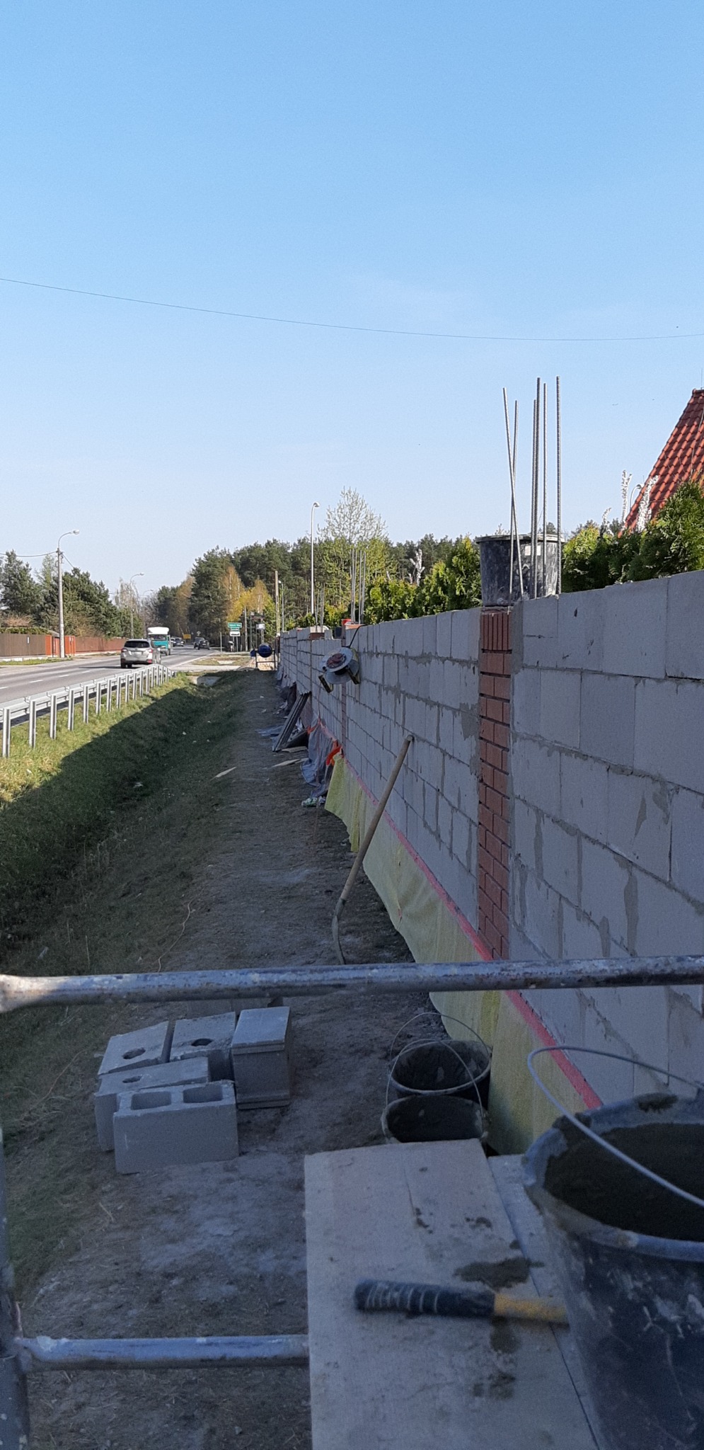 Wznoszenie ogrodzenia z bloczków betonowych z fragmentami cegły klinkierowej, widoczne narzędzia murarskie i zaprawa, w tle droga i zieleń.