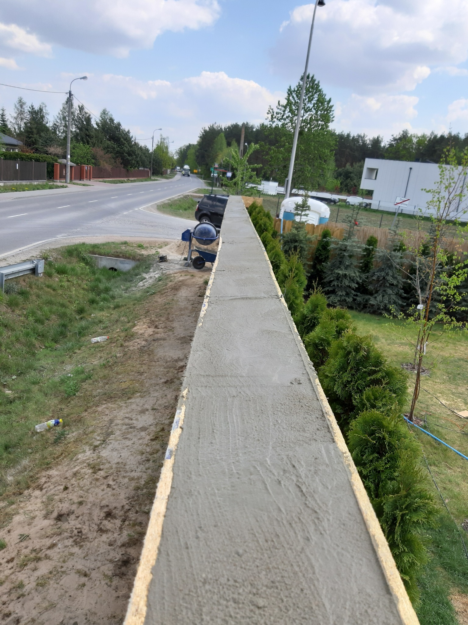 Świeżo wylany betonowy murek oporowy z drewnianym szalunkiem, widoczna betoniarka i droga w tle.