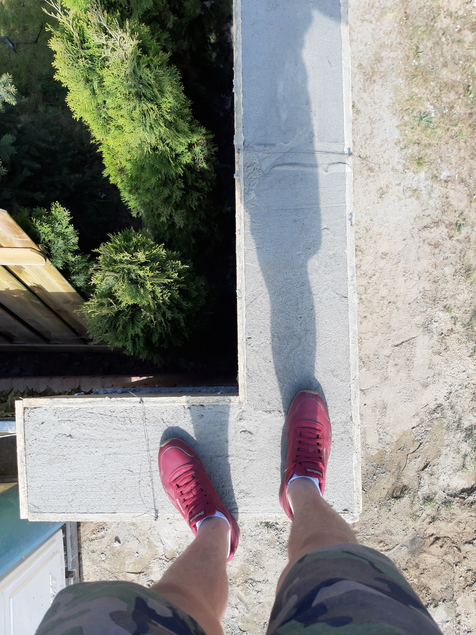 Widok z góry na świeżo wylany betonowy fundament pod ogrodzenie, z widocznymi czerwonymi butami i nogami w krótkich spodenkach moro, oraz tujami w tle.