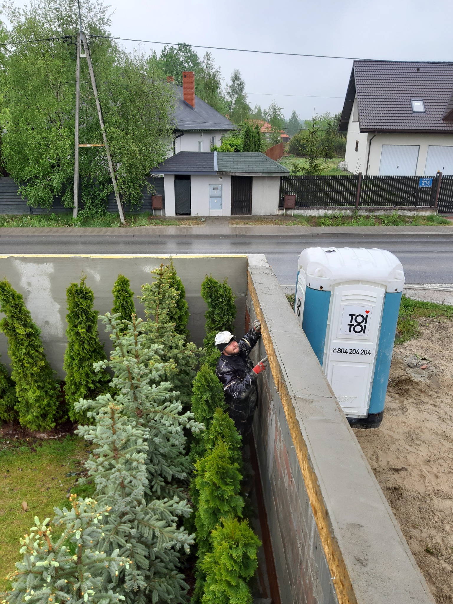 Pracownik w roboczym ubraniu, w trakcie wylewania betonu na mur oporowy, widoczna przenośna toaleta Toi Toi, oraz rząd tujek i świerków posadzonych wzdłuż muru.
