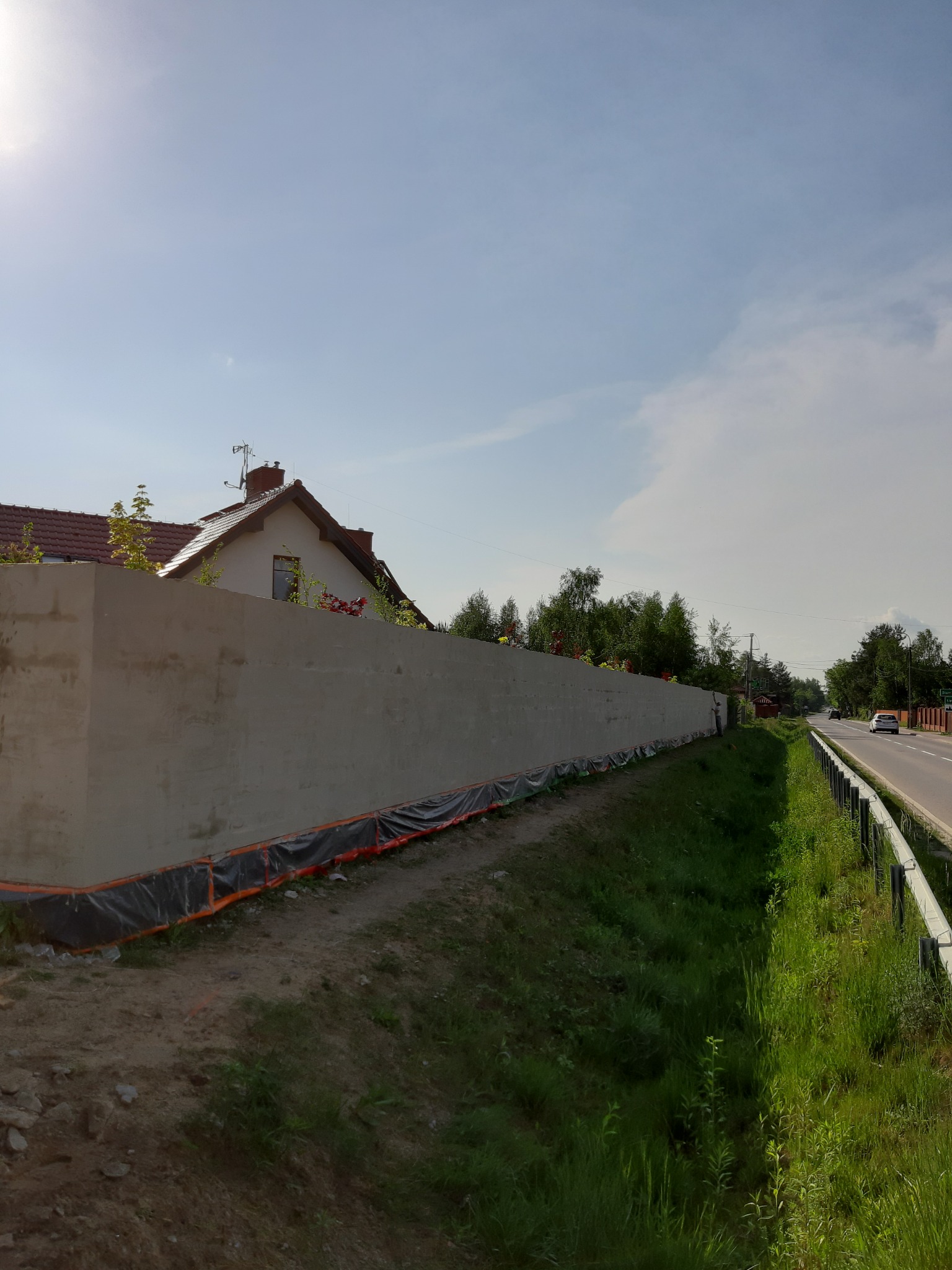 Długi, świeżo otynkowany mur oddzielający posesję z domem od ruchliwej drogi, zabezpieczony folią ochronną u podstawy.