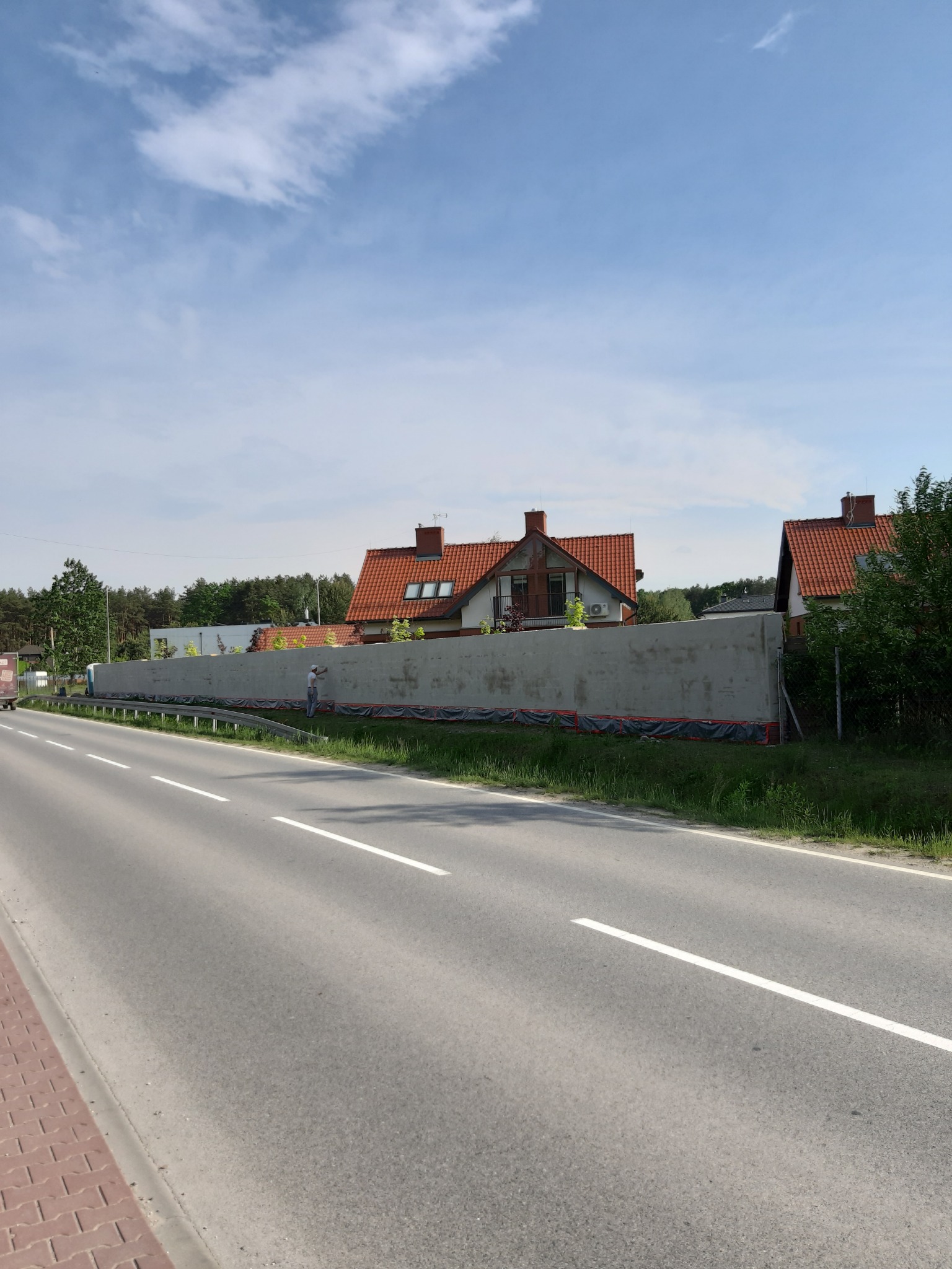 Długi, świeżo otynkowany mur oddzielający posesję od drogi, widoczny pracownik w jasnym ubraniu wykonujący drobne poprawki, w tle domy z czerwoną dachówką i błękitne niebo.
