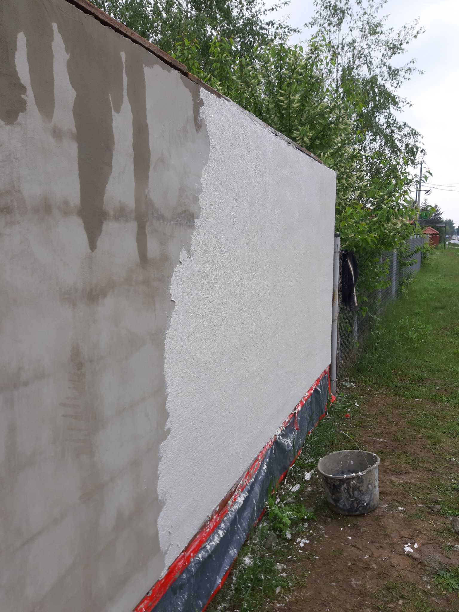 Ściana budynku w trakcie nakładania tynku, widoczne fragmenty surowego i świeżo nałożonego tynku, kubeł z zaprawą przy ścianie, zabezpieczenie folią u podstawy ściany.