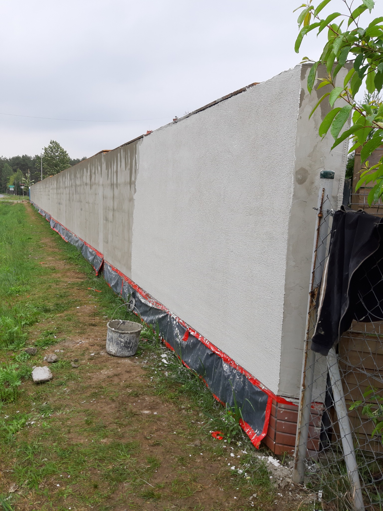 Długi, otynkowany mur ogrodzeniowy w trakcie prac, częściowo mokry od świeżo nałożonego tynku, zabezpieczony folią u podstawy, widoczny kubeł i resztki materiałów na trawie.