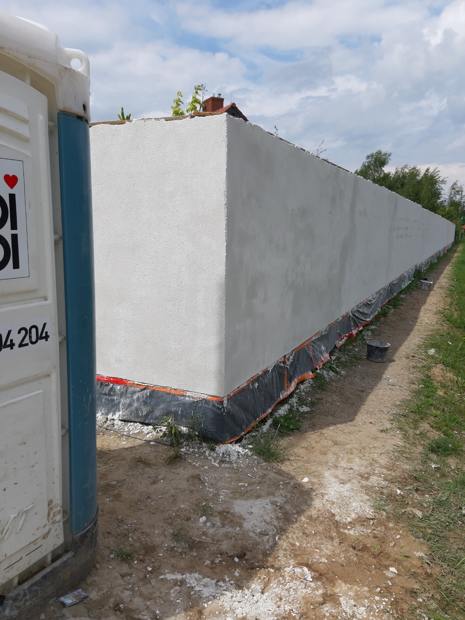 Długi, świeżo otynkowany mur z widoczną strukturą tynku, zabezpieczony folią u podstawy, z przenośną toaletą w lewym dolnym rogu i rozrzuconymi fragmentami gruzu na ziemi.