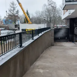 Czarna metalowa balustrada zamontowana na betonowym murku oporowym wzdłuż podjazdu do garażu w nowoczesnym budynku z cegły klinkierowej, w tle plac budowy i żuraw.