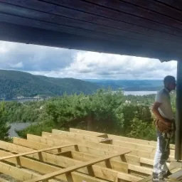 Konstrukcja drewnianego tarasu w trakcie budowy, z widokiem na zalesione wzgórza i jezioro w tle, mężczyzna z narzędziami przy kolumnie, ceglana ściana po prawej stronie.