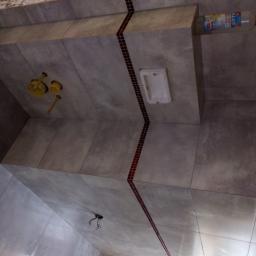 Przemysław glowacki - Wykończona łazienka z szarymi płytkami, dekoracyjnym pasem z małych czerwonych płytek, widoczne elementy instalacji sanitarnej i elektrycznej, oraz butelka z płynem na półce.