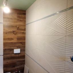Przemysław glowacki - Łazienka w trakcie remontu: ściana wyłożona płytkami z falistym wzorem w odcieniach beżu, obok fragment ściany z panelami imitującymi drewno. Widoczne elementy instalacji sanitarnej i surowy sufit.