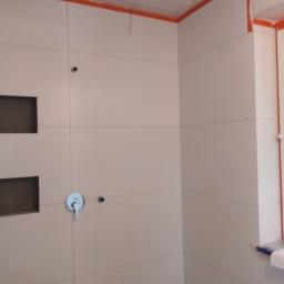 Przemysław glowacki - Ściana łazienki w trakcie wykańczania w Ciechanowie, z widocznymi jasnymi płytkami, wnękami, baterią umywalkową i oknem zabezpieczonym taśmą malarską.