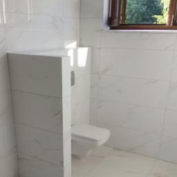 Przemysław glowacki - Wykończona łazienka z podwieszaną toaletą w zabudowie, białe płytki z delikatnym marmurkowym wzorem na ścianach i podłodze, drewniane okno z widokiem na zieleń na zewnątrz.