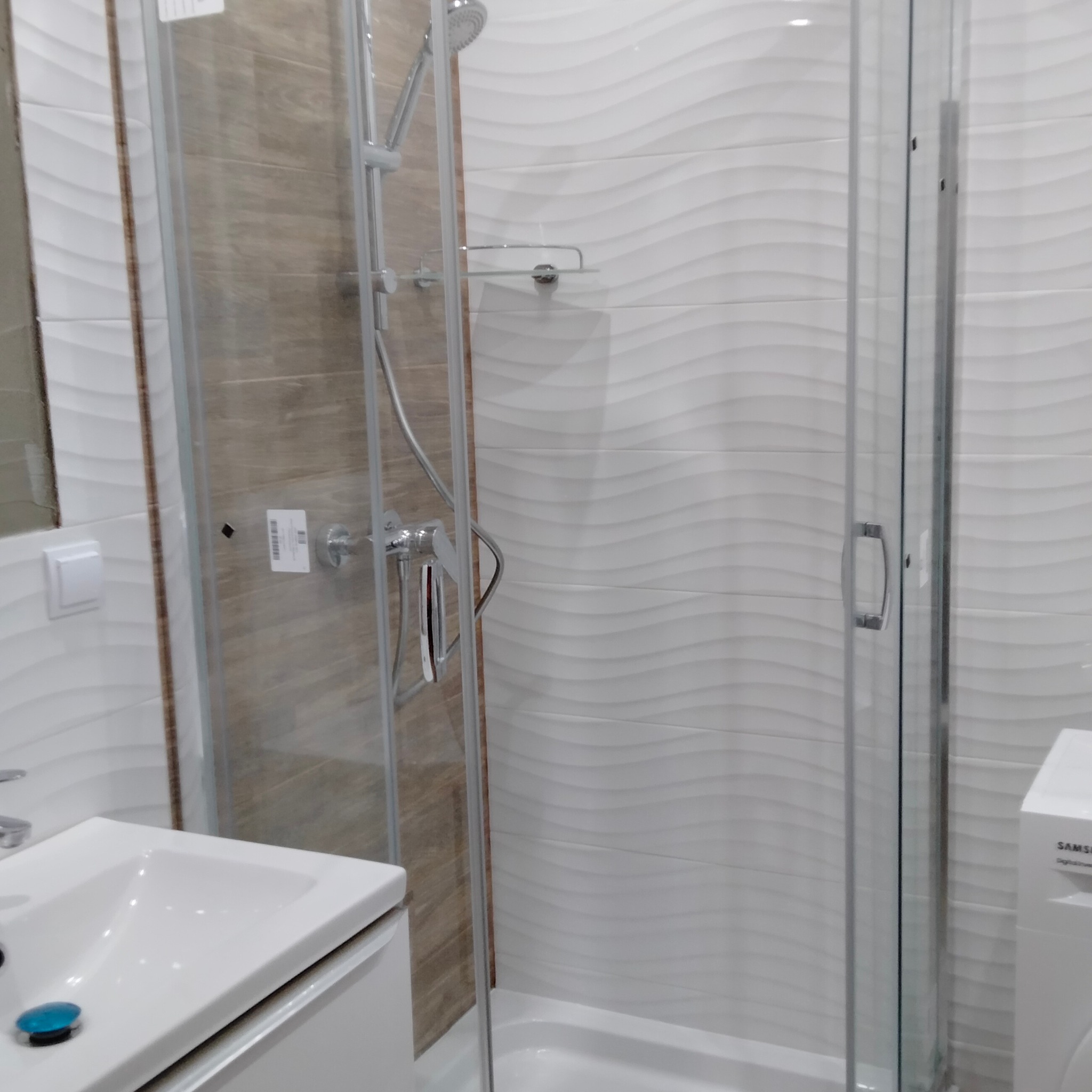 Nowoczesna łazienka z kabiną prysznicową, białą umywalką i pralką, wykończona płytkami w odcieniach bieli i beżu z subtelnym wzorem fal.