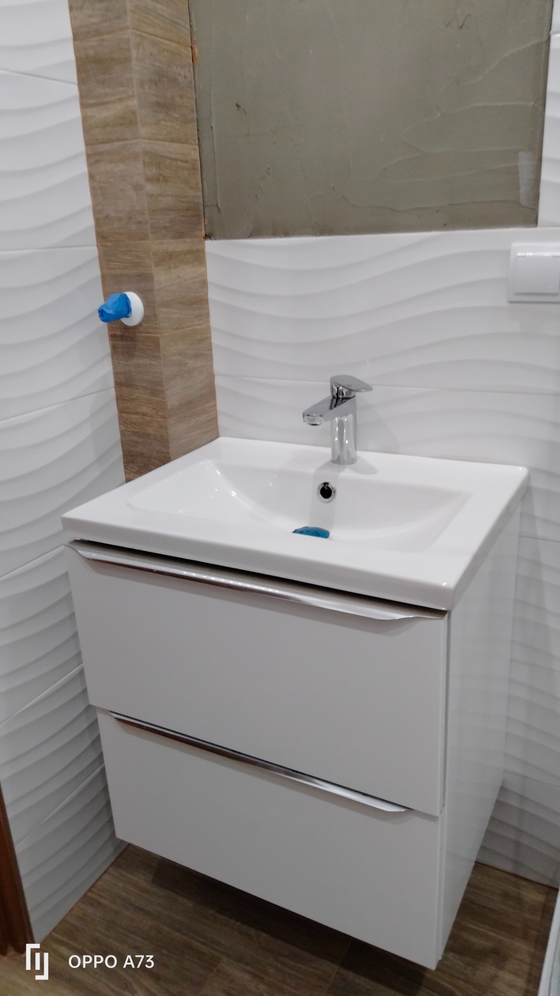 Nowoczesna, biała szafka pod umywalkę z chromowanymi uchwytami, na tle ściany z dekoracyjnymi, falistymi płytkami i fragmentem ściany w trakcie kładzenia płytek.