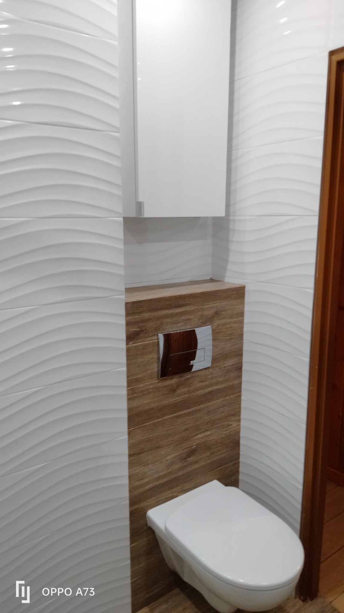 Nowoczesna toaleta podwieszana z chromowanym przyciskiem spłukującym, obudowana płytkami imitującymi drewno, na tle białych, falistych płytek ściennych i wiszącej szafki.