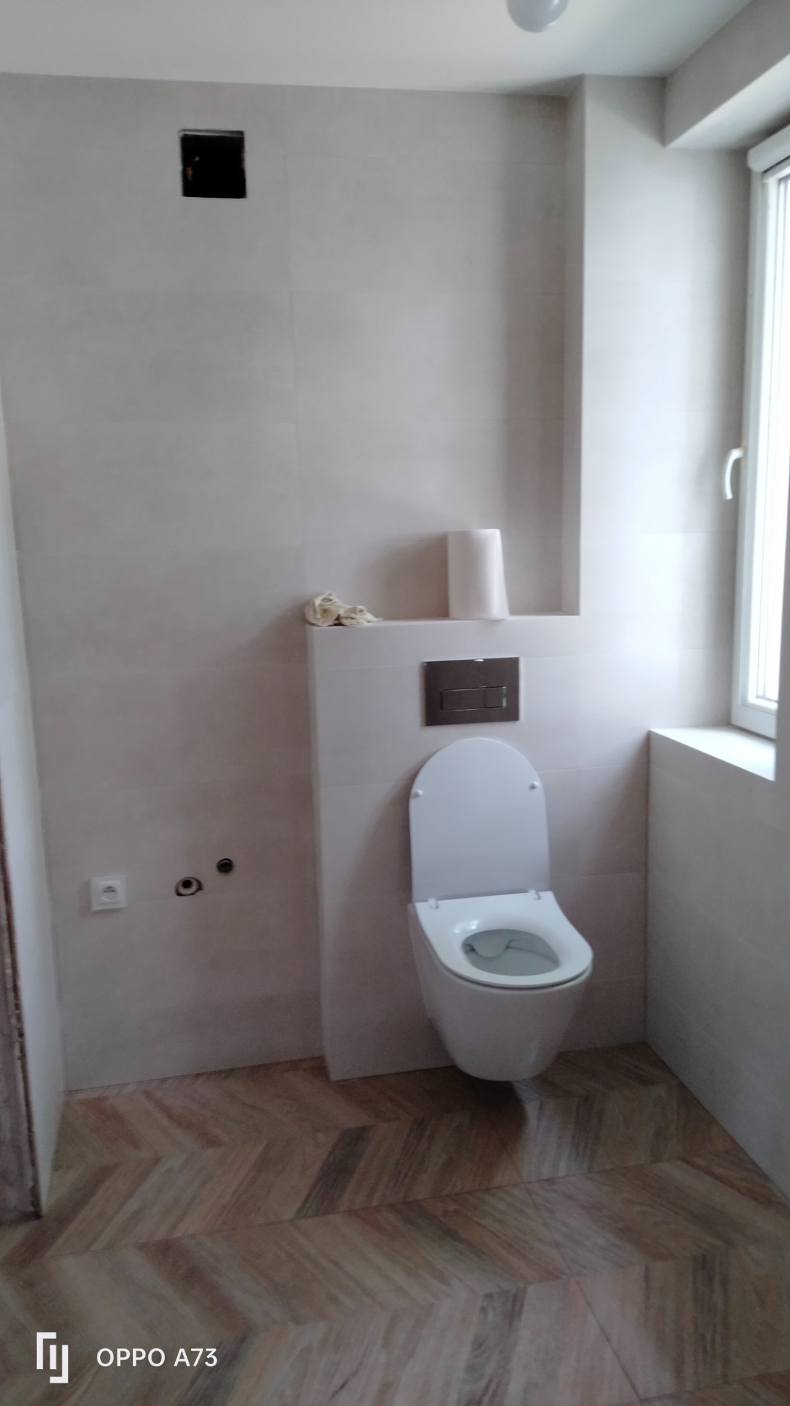 Nowoczesna, biała toaleta podwieszana z chromowanym przyciskiem spłukującym, zamontowana na tle jasnych, kwadratowych płytek ściennych i podłogi ułożonej we wzór jodełki, z widocznymi...