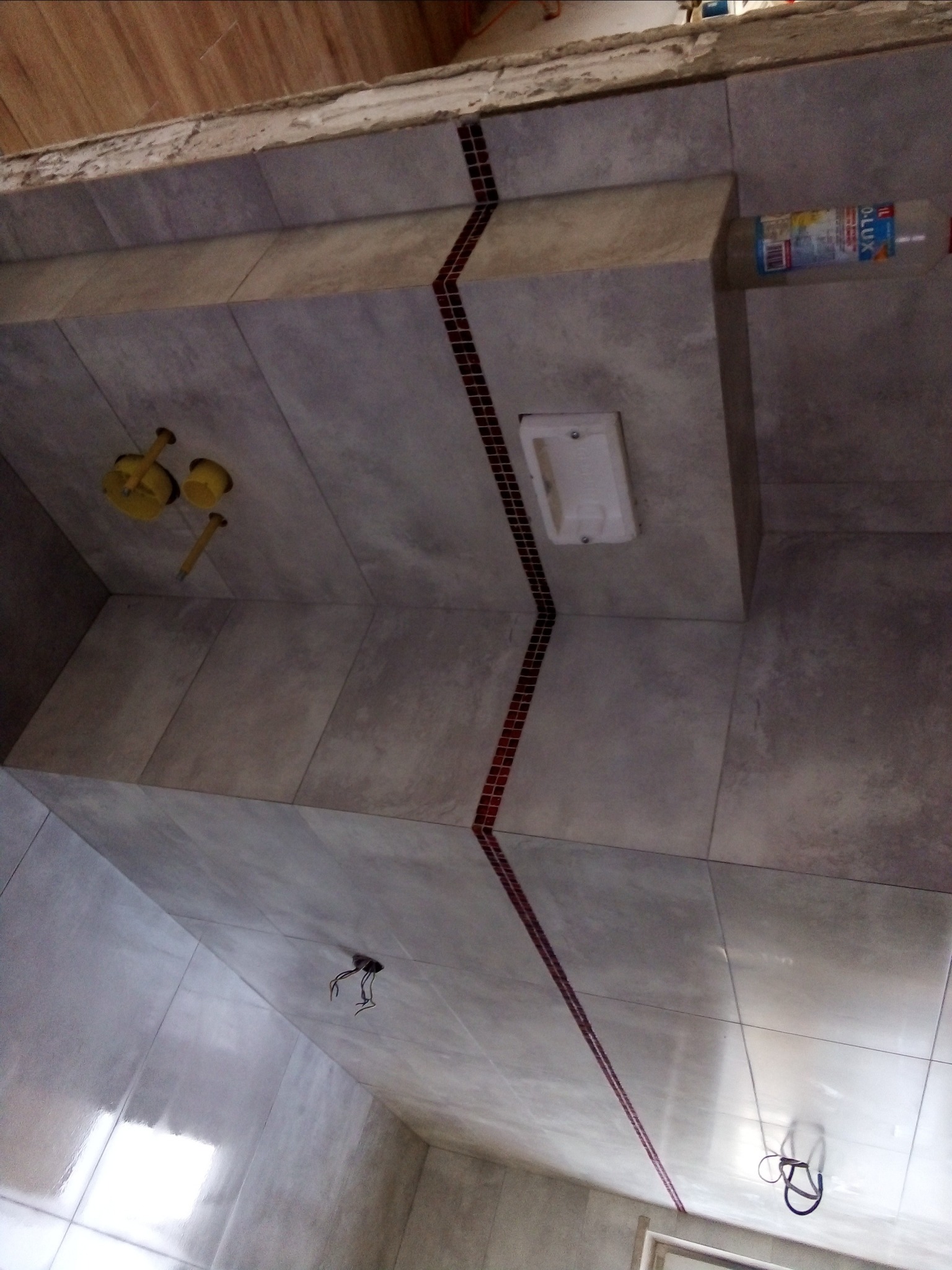 Wykończona łazienka z szarymi płytkami, dekoracyjnym pasem z małych czerwonych płytek, widoczne elementy instalacji sanitarnej i elektrycznej, oraz butelka z płynem na półce.