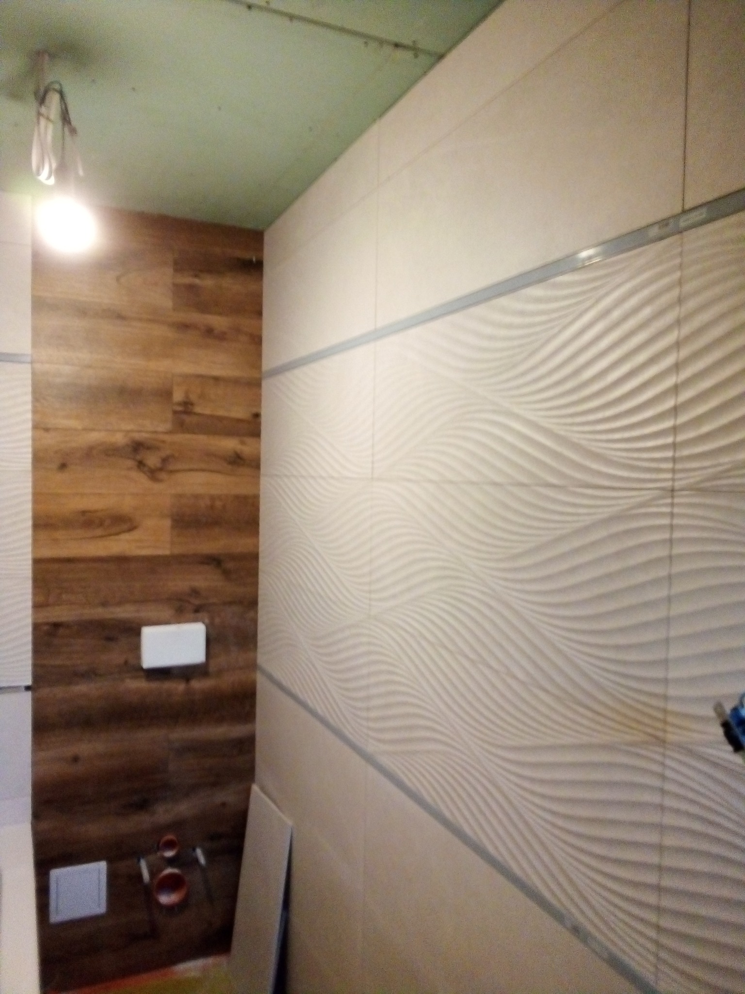 Łazienka w trakcie remontu: ściana wyłożona płytkami z falistym wzorem w odcieniach beżu, obok fragment ściany z panelami imitującymi drewno. Widoczne elementy instalacji sanitarnej i surowy sufit.