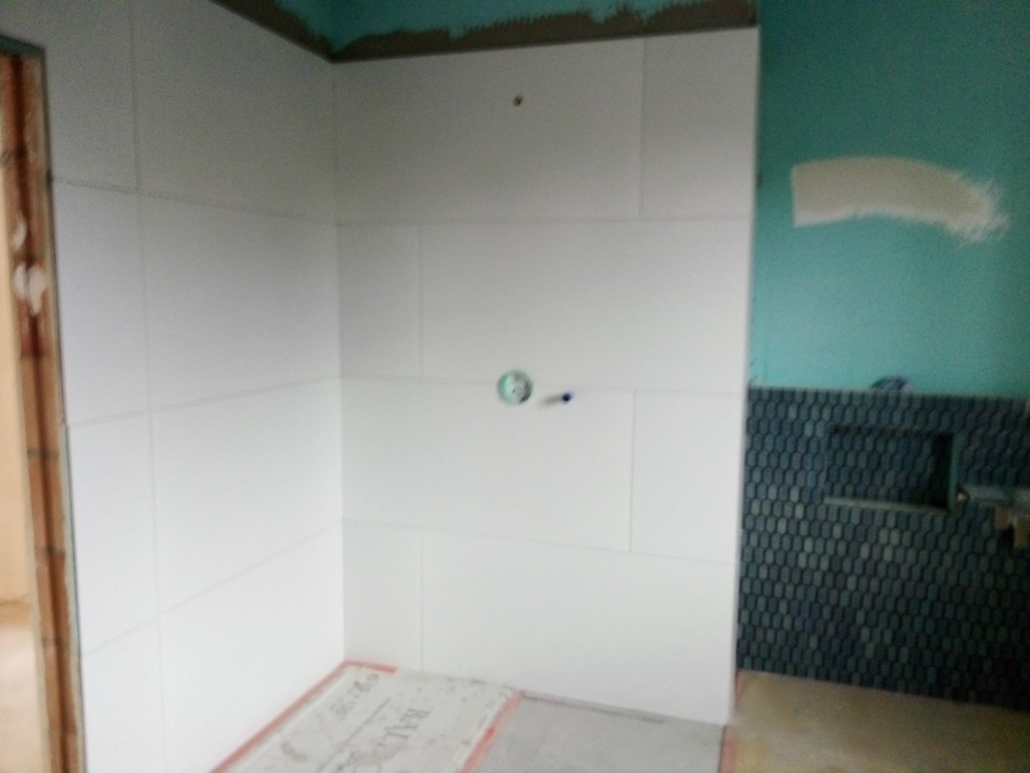 Wnętrze łazienki w trakcie wykańczania: białe, prostokątne płytki ścienne częściowo ułożone, widoczne ślady zaprawy na ścianie, fragment ściany z mozaiką w odcieniach szarości, przygotowane miejsce...