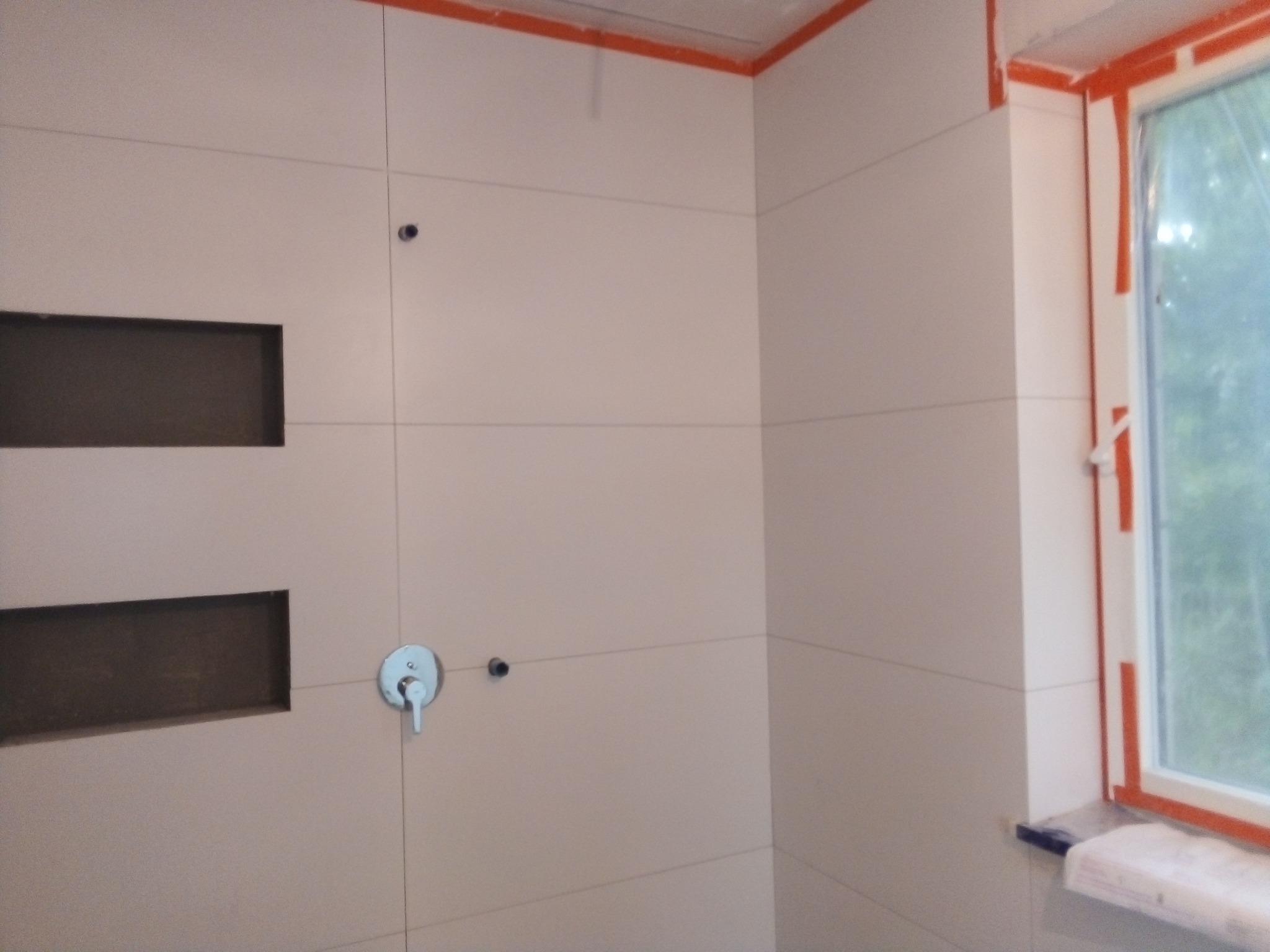 Ściana łazienki w trakcie wykańczania w Ciechanowie, z widocznymi jasnymi płytkami, wnękami, baterią umywalkową i oknem zabezpieczonym taśmą malarską.